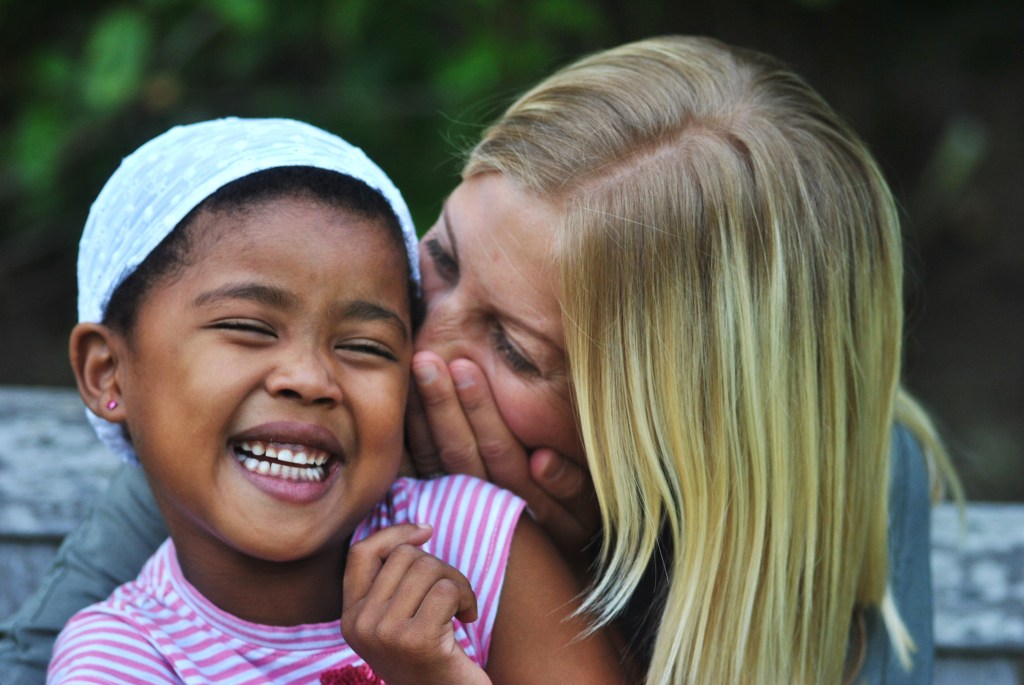 Mother whispering into daughters ear and laughing together on a bench outside