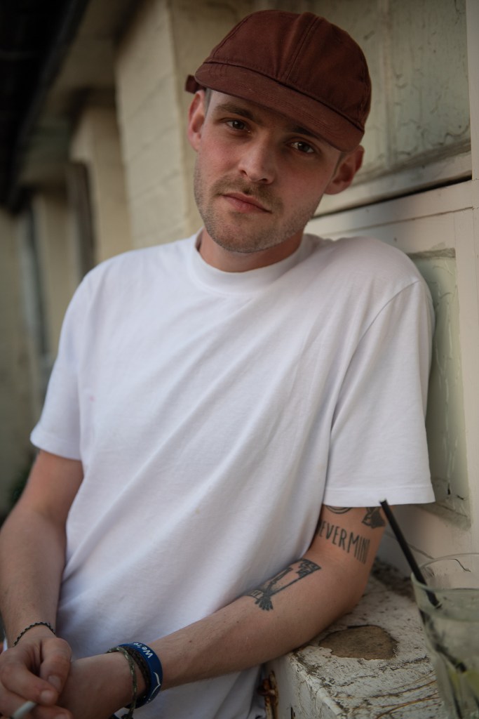 Portrait of a young man with tattooed arms leaning against a brick wall, wearing a burgundy red cap and white t shirt 