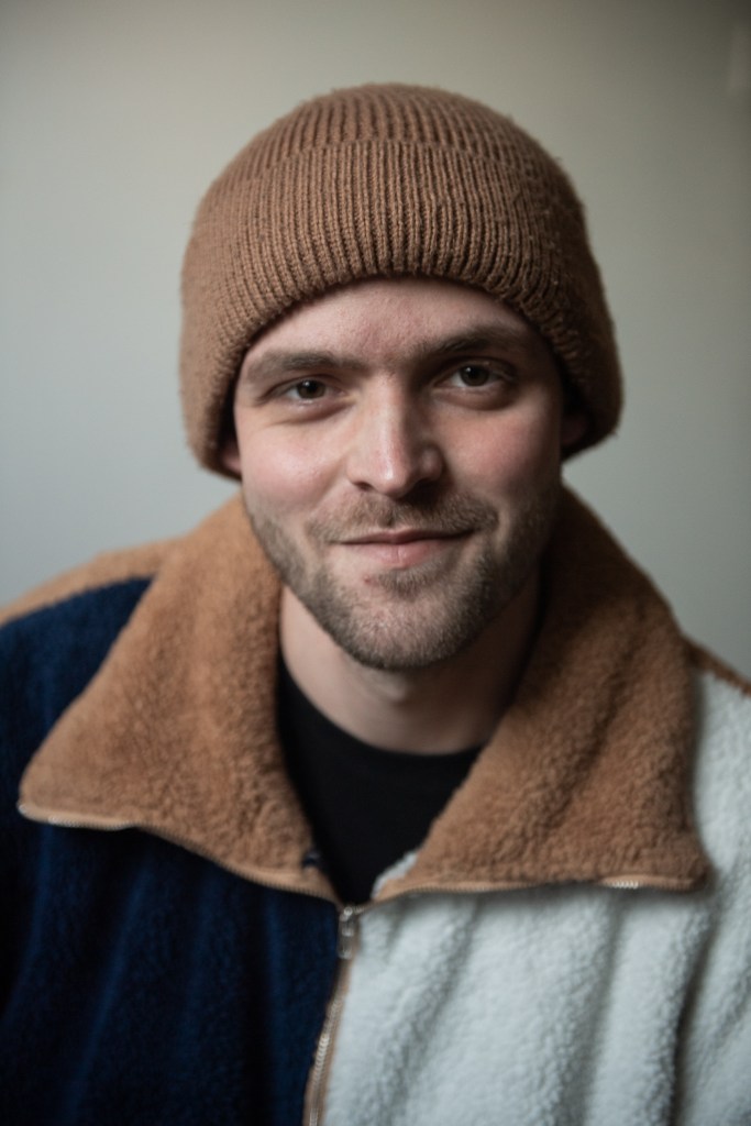 Portrait of a man smiling looking directly into camera, with brown hat and textured jumper.