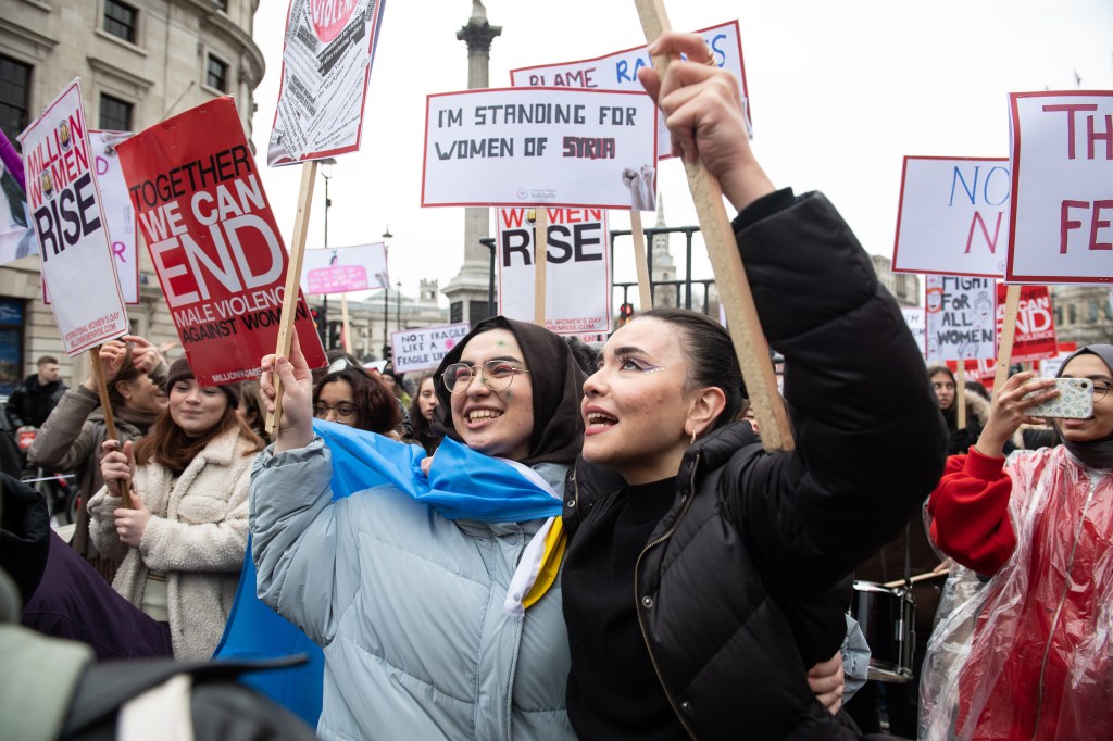 Two young women holding protest signs with arms round each other, smile and look up during the Million Women Rise protest 