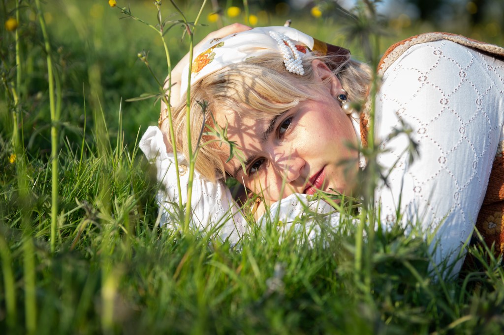 Portrait of a young woman laying in the grass surrounded by buttercup flowers, with a headscarf and white lace shirt 