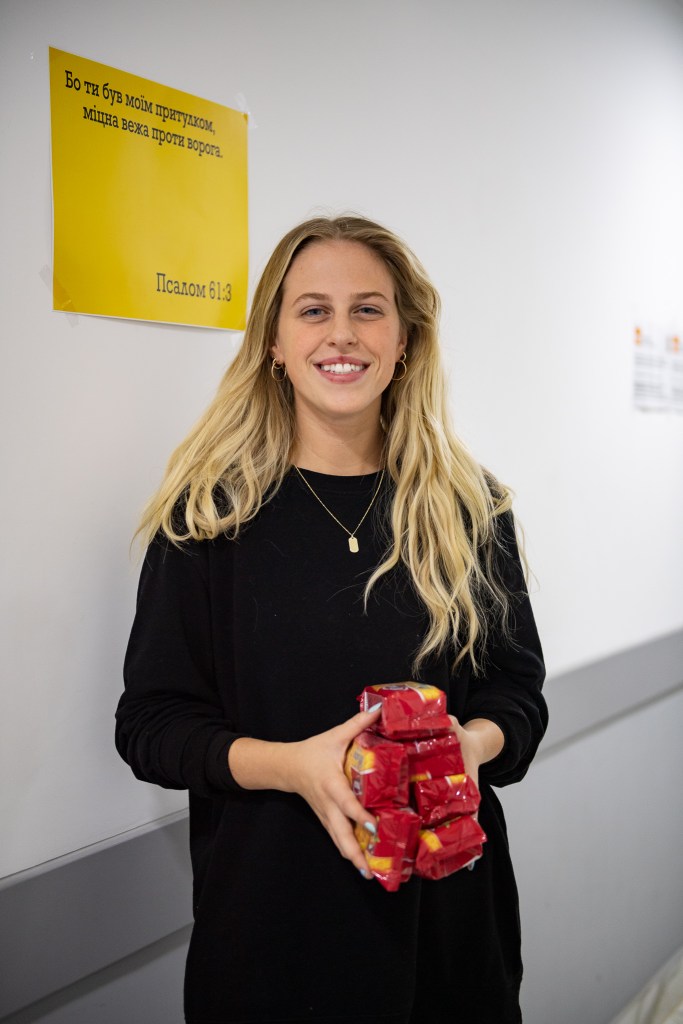 A young volunteer in a corridor of a refugee centre smiling, holding food for the families staying there.