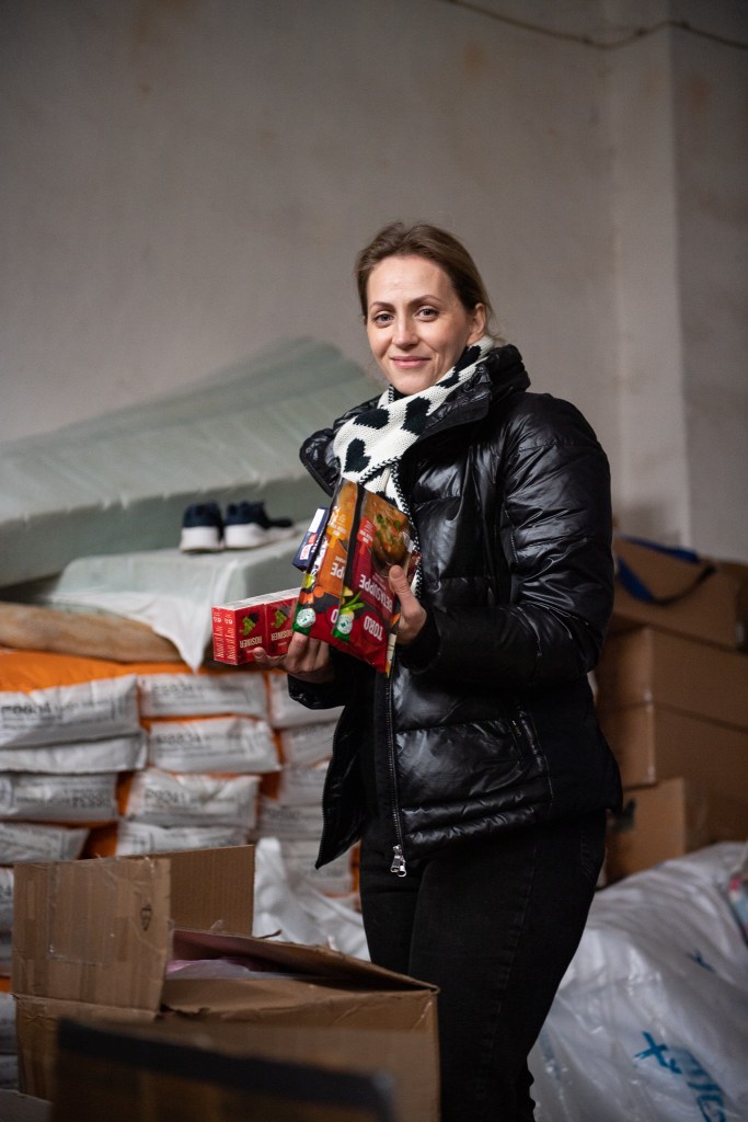 A volunteer in a warehouse of a refugee centre smiling, finding food for an an expectant family staying at the camp.