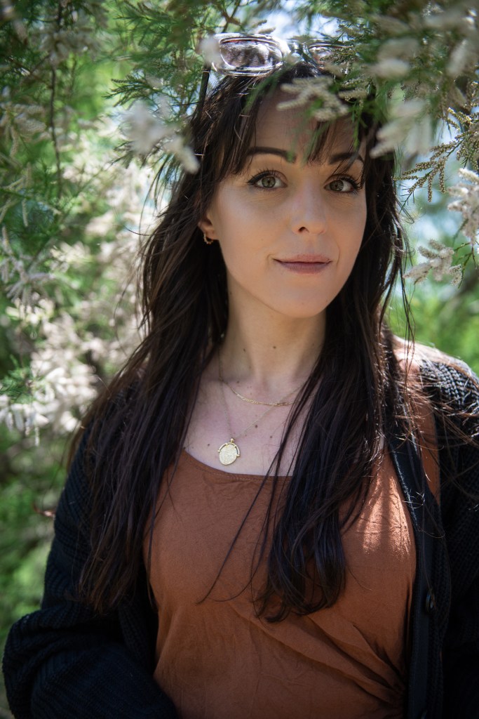 Woman with sunglasses smiles surrounded by white flowers and green foliage 